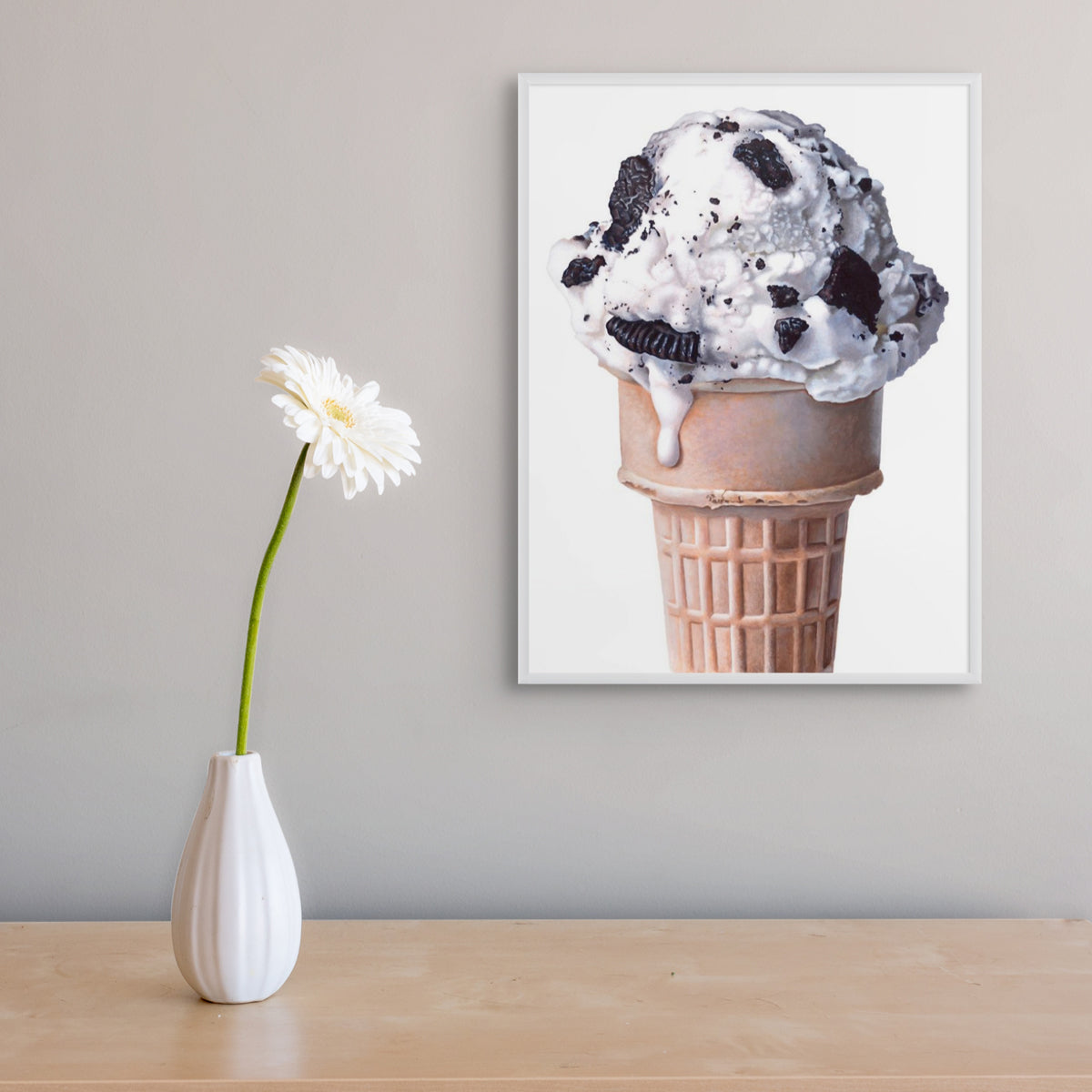 Cookies and cream ice cream cone art print in a frame on a wall next to a table with a flower in a vase.